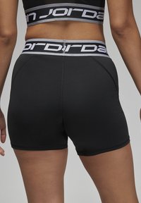 Crne atletske kratke hlače izrađene od glatkog materijala s širokim sivo-belim pojasom na kojem se nalazi "Jordan" logo u crnom slovu.