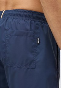 Tengerészkék fürdőnadrág elasztikus derékpánttal, oldalzsebbel és egy kis "BOSS" logó címkével. Simító, könnyű anyag.