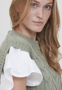 Strickpullover in hellgrün mit einem strukturierten Muster, über einem weißen rüschenshirt mit kurzen Ärmeln getragen; Nahaufnahme mit Fokus auf die Stoffdetails.