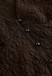 Tissu en dentelle brun foncé avec des motifs circulaires texturés et des boutons ronds brillants disposés verticalement.