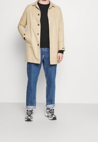 Giacca di cotone beige con quattro bottoni, tasca sul petto e tasche laterali, indossata su una camicia nera. Abbinata a jeans blu e sneakers grigie.