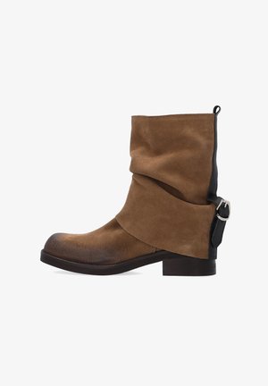 Brązowy zamszowy botek do kostki o luźnym kroju, zaokrąglonym nosku, czarnych skórzanych akcentach oraz szczególe z klamrą z boku. Niski, stworzony z jednego kawałka obcas.