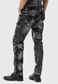 Jeans en camouflage noir et gris avec une coupe slim, dotés de plusieurs poches et d'un tissu texturé. Ceinture noire avec des clous.