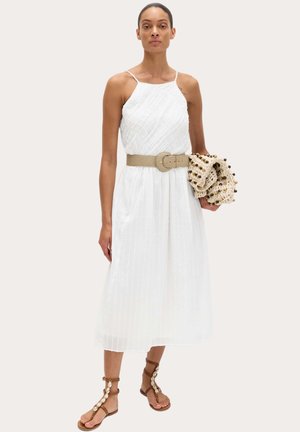Kvinde iført en hvid, ærmeløs midi-kjole med et beige bælte, der holder en beige tekstureret clutch og bærer brune sandaler med skaldetaljer.