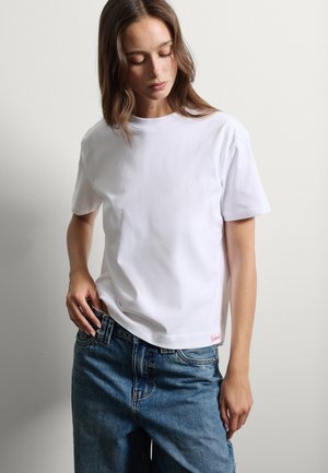 T-shirt bianco a maniche corte in cotone, con scollo rotondo e taglio dritto. Logo del marchio rosso in basso. Indossato con jeans in denim blu.