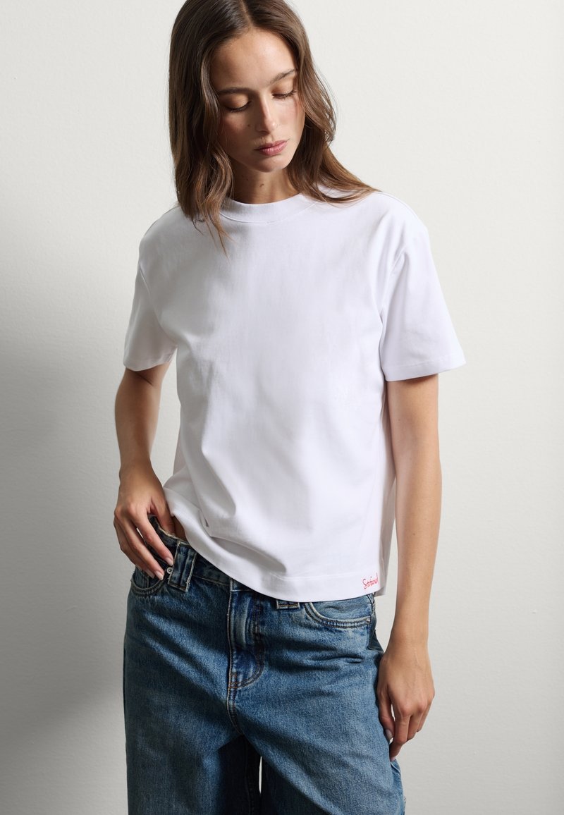 T-shirt bianco a maniche corte in cotone, con scollo rotondo e taglio dritto. Logo del marchio rosso in basso. Indossato con jeans in denim blu.