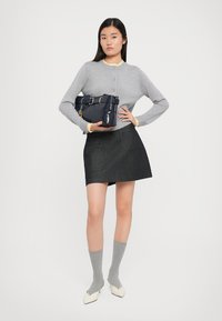 Cardigan grigio con rifiniture gialle, gonna corta scura, calzini grigi e scarpe ballerine beige a punta, che tiene una borsa blu navy con dettaglio a cintura.