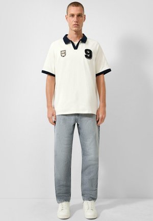 Mann trägt weißes Polohemd mit schwarzem Kragen und Nummer-neun-Patch, hell gewaschene Jeans und weiße Turnschuhe, steht vor einfachem Hintergrund.