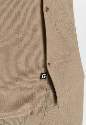 Gros plan sur un tissu côtelé beige avec rabat boutonné et petite étiquette noire portant un logo "r" stylisé en blanc.