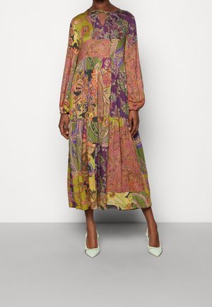 Nainen, joka pukeutunut pitkään, väljästi istuvaan mekkoon, jossa on monivärinen paisley-kuvio ja pitkät hihat, seisoo vaaleanvihreissä korkokengissä.