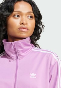Fialová mikina na zips vyrobená z mäkkého materiálu, s vysokým golierom, tromi bielymi pruhmi na pleciach a bielym logom Adidas na hrudi.
