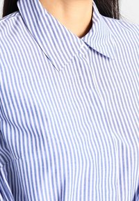 Niebiesko-biała koszula w paski z ostrym kołnierzykiem, z cienkimi pionowymi liniami i gładką fakturą. Widoczne zapięcie na guziki z przodu.