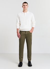 Un pull en maille blanche avec un col, associé à un pantalon taper olive et des chaussures noires, se tenant contre un fond uni.