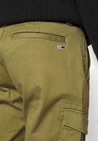 Gros plan d’un pantalon cargo vert olive avec poche arrière et petit patch logo rectangulaire rouge, blanc et bleu sur le côté droit.