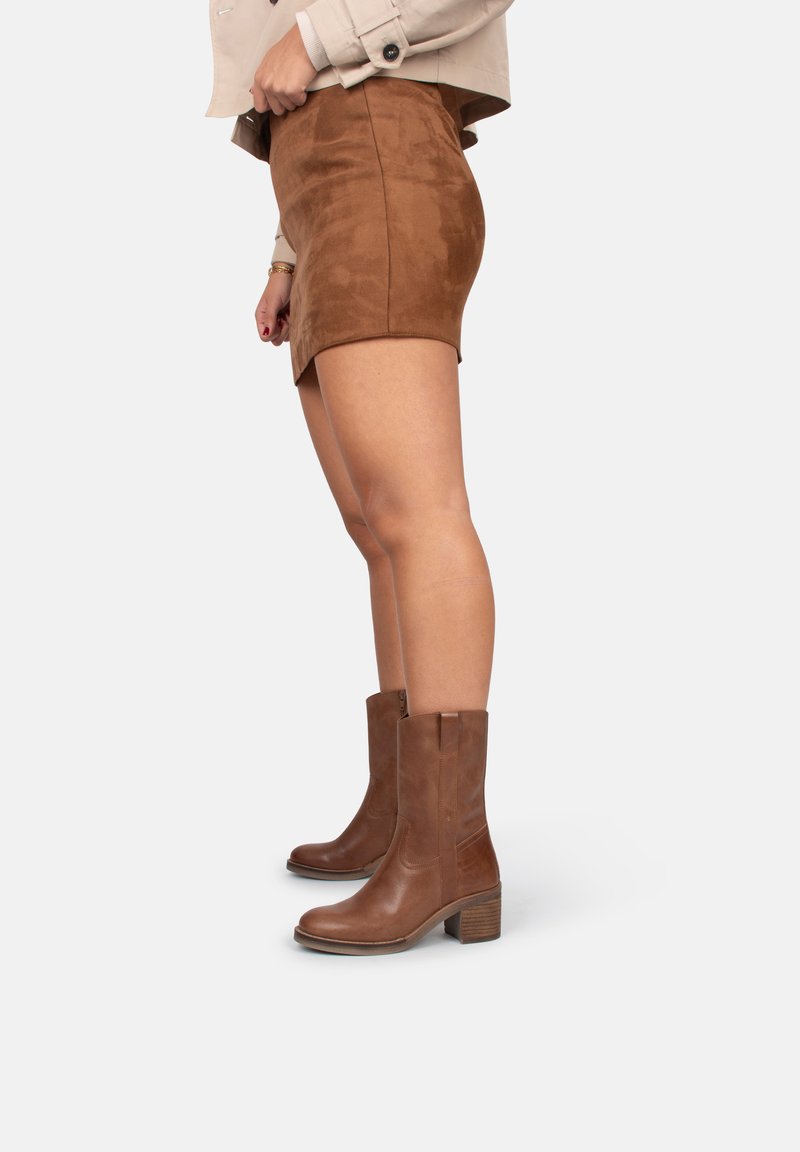 Botas de cuero marrón hasta el tobillo con tacón cuadrado, superficie texturizada y cremalleras laterales, combinadas con una minifalda de ante marrón.