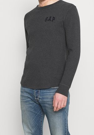 Szary longsleeve z prążkowanym wzorem, okrągłym dekoltem i ciemnym napisem "GAP" na piersi, w zestawieniu z niebieskimi dżinsami.