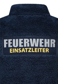 Dunkelblaue Fleecejacke aus einem strukturierten, flauschigen Material. Großer Aufnäher mit "FEUERWEHR" in Grau und "EINSATZLEITER" in Gelb.