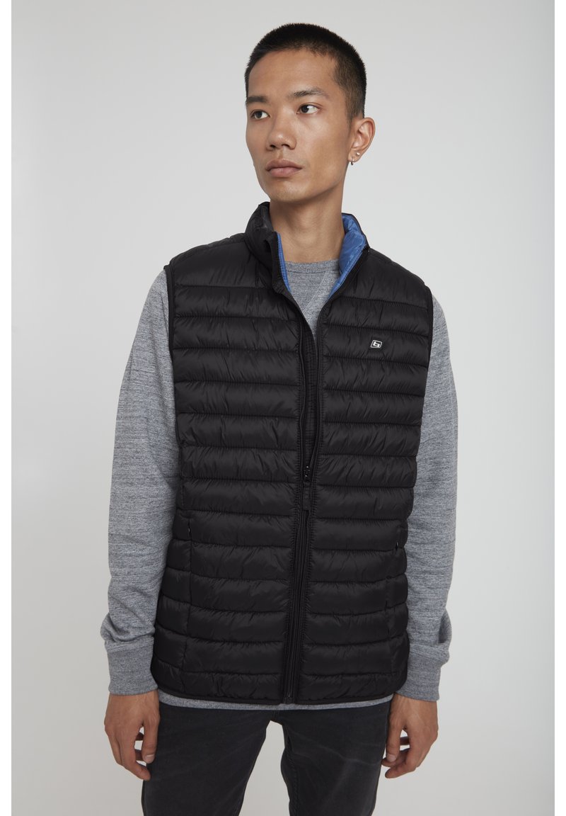 Schwarze, gepolsterte Weste mit durchgehendem Reißverschluss, einem blauen Innenfutter und einem kleinen Logo. Getragen über einem grauen Langarmshirt.