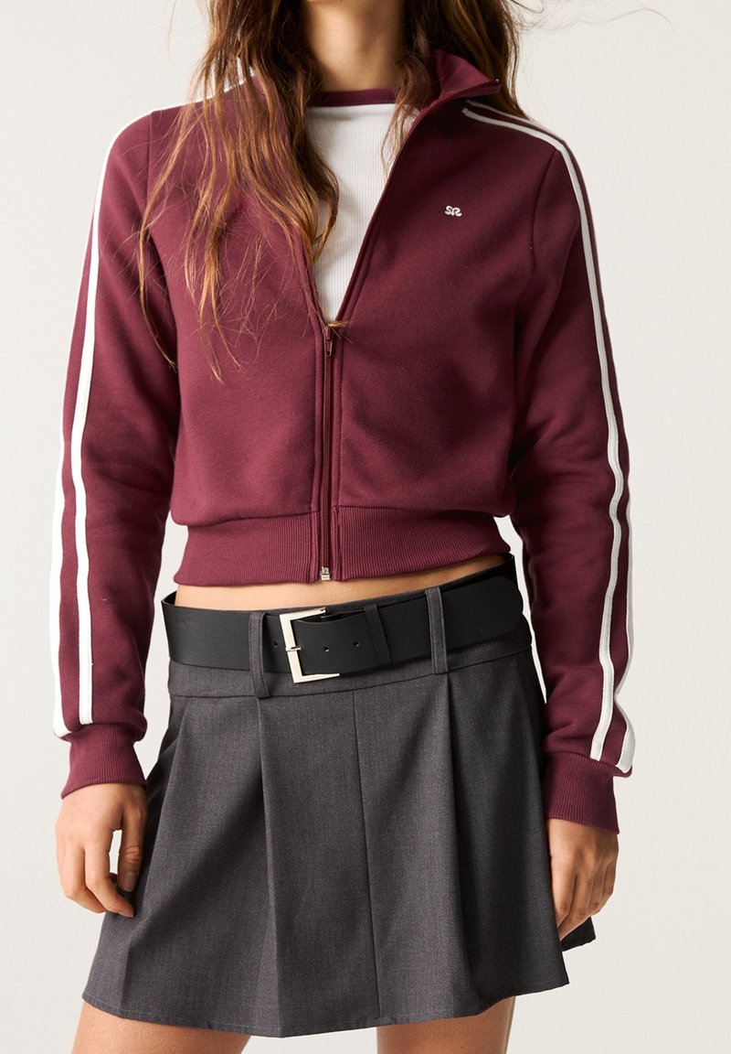 Femme portant une veste zippée courte bordeaux avec des rayures blanches sur les manches, une chemise blanche en dessous, et une jupe plissée grise avec une ceinture noire.