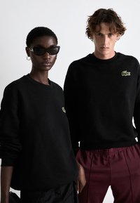 Crni duksevi sa zelenim Lacoste krokodil logotipom, upareni sa crnim naočalama. Muški model ima tamnu, valovitu kosu i nosi bordo hlače.