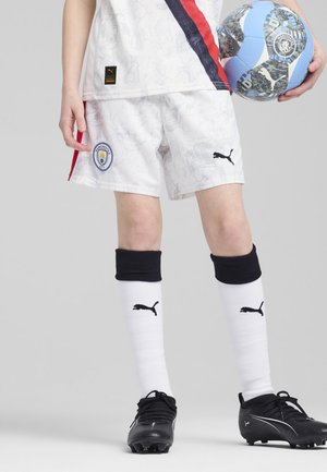 Ungdomsspelare i fotboll med mönstrade vita shorts med Manchester City-logotyp, svarta strumpor och svarta fotbollsskor, håller en blå och svart fotboll.