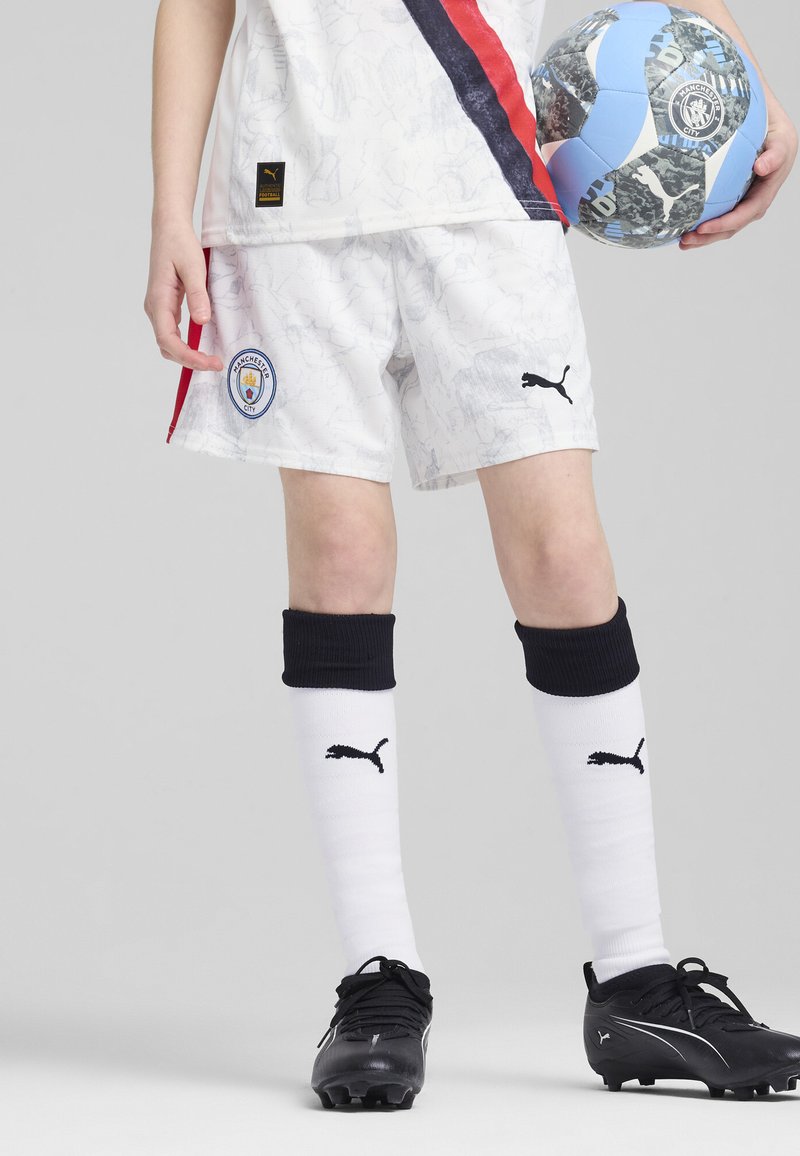 Jugador de fútbol juvenil con pantalones cortos blancos estampados con el logo del Manchester City, calcetas negras y botas negras, sosteniendo un balón de fútbol azul y negro.