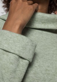Pull vert clair en tissu doux et texturé. Présente un col haut plié et des poignets roulés. Vue en gros plan de la main et des détails du tissu.