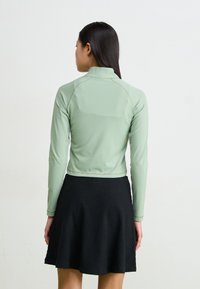 Zaļš, garām piedurknēm, kropts tops no gludas auduma, apvienots ar melnu ribotu svārku. Dizainā ir iekšējā apkakle un cieši pieguļošas piedurknes.