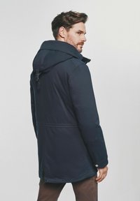 Mężczyzna z brodą ubrany w granatowy płaszcz z kapturem i brązowe spodnie, zwrócony bokiem wobec jasnego, jednolitego tła.