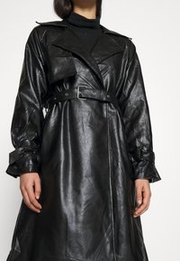 Czarny skórzany płaszcz typu trench z paskiem w talii, szerokimi klapami i zawiązywanymi mankietami. Gładka tekstura i elegancki fason.