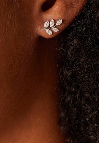 Silberne, blattförmige Ohrringe mit mehreren klaren Kristallen, die ein geschwungenes Design und eine strukturierte Oberfläche aufweisen, vor einem dunkelhäutigen Teint und lockigem Haar.