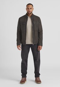 Dunkle Lederjacke mit Stehkragen, zwei Brusttaschen, kombiniert mit einem beigen Pullover, dunklen Jeans und braunen Wanderschuhen.