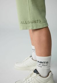 Lys grønne shorts med "ALLSAINTS" broderet, parret med hvide teksturerede sokker med sort skrift og hvide sneakers.