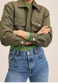 Olivgrüne Zip-Jacke mit zwei Brusttaschen, silbernen Druckknöpfen und weiten Ärmeln, über einem gemusterten grünen Oberteil und blauen Jeans.