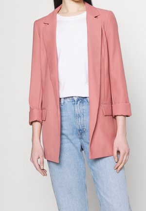 Kvinde iført lyserød blazer med opkørte ærmer over hvid skjorte kombineret med lyseblå højtaljede jeans, stående mod en ensfarvet baggrund.