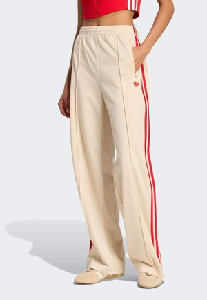 Beige wide-leg track pants with red side stripes, elastic waistband, zippered side pocket, and small red logo near pocket.