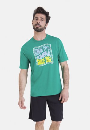 Homme aux cheveux foncés et barbe portant un T-shirt vert avec l'inscription "Urban Style Athena Since 1962" et un short noir, debout devant un fond uni.