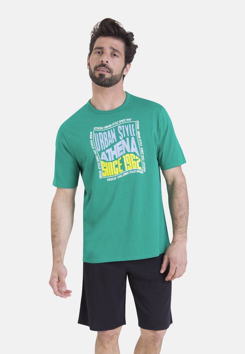 Homme aux cheveux foncés et barbe portant un T-shirt vert avec l'inscription "Urban Style Athena Since 1962" et un short noir, debout devant un fond uni.