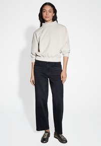 Felpa beige chiaro con collo alto e cintura elastica, abbinata a jeans neri a gamba dritta e scarpe nere a profilo basso.