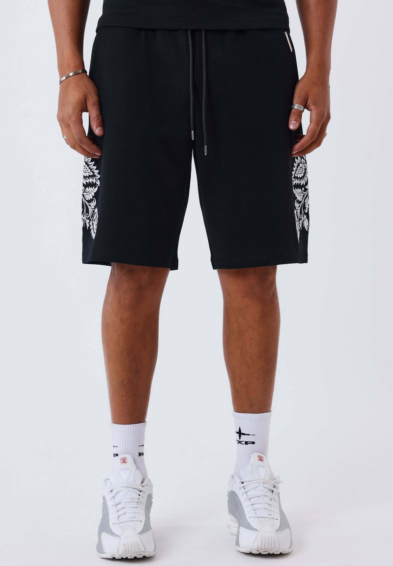 Uomo che indossa pantaloncini neri con motivi floreali bianchi sui lati, calzini bianchi e scarpe da ginnastica bianche, in piedi dritto con le mani rilassate.