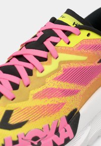 Gros plan sur une chaussure de course jaune et rose avec des lacets roses, une languette noire et une semelle blanche arborant un logo de marque visible.