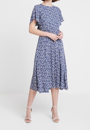 Vrouw draagt een blauw midi-jurk met witte bloemenprint, korte vleermuismouwen en witte hoge hakken met enkellus.
