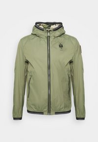 Chaqueta ligera de color verde oliva con capucha, cierre frontal con cremallera, detalles de ribete negro, bolsillos laterales y forro de camuflaje.