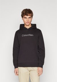 Sweat à capuche noir avec une poche kangourou à l'avant, présentant un logo Calvin Klein blanc dans une police texturée. Tissu doux avec des poignets et un ourlet côtelés.