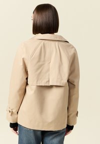 Beżowy płaszcz trench z wyraźnie zarysowanym kołnierzem, skróconym tyłem i guzikiem na mankietach. Gładka faktura, o luźnym kroju i minimalistycznym designie.