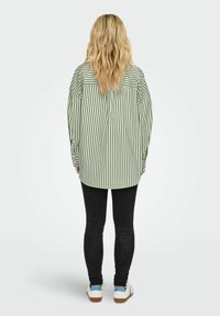 Groen en wit gestreepte oversized blouse met lange mouwen, voorzien van een plooi aan de achterkant en zijzakken, gecombineerd met zwarte skinny jeans en witte sneakers.