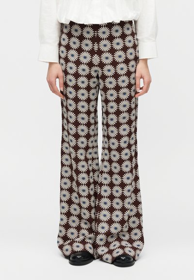 Καφέ παντελόνια με φαρδιά γραμμή και λευκά σχέδια μαργαρίτες με μπλε κέντρα, συνδυασμένα με λευκό μακρυμάνικο πουκάμισο και μαύρα παπούτσια.