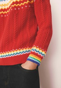 Rode gebreide trui met een gestructureerd kabelontwerp, kleurrijke zigzagpatronen bij de schouderpas en mouwen, met accentkleuren in regenboogstrepen.