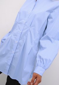 Ljust blå skjorta med vita vertikala ränder, långa puffärmar, rundad nertill och en avslappnad passform. Tillverkad av ett glatt tyg.