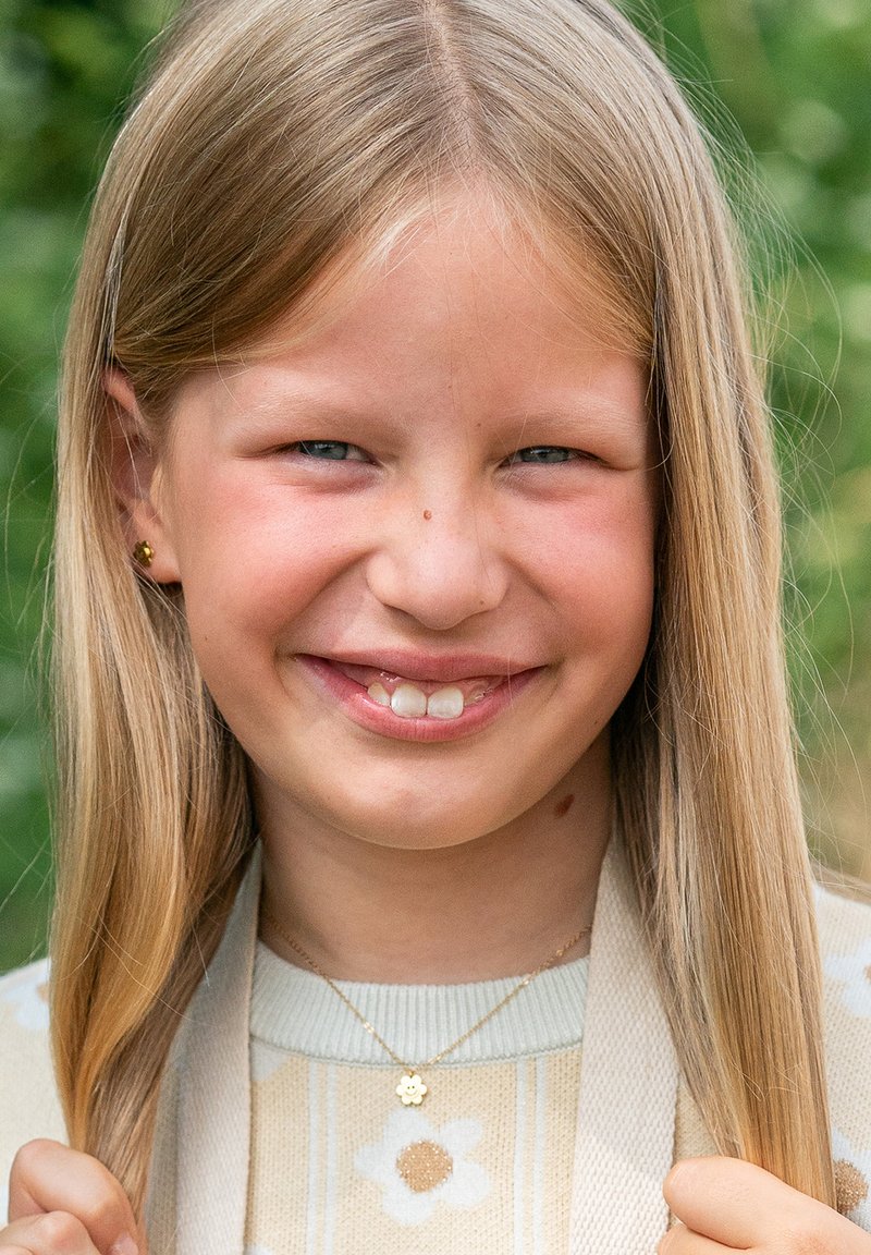 Fille blonde avec de longs cheveux, portant un pull crème à motifs floraux et des bijoux en or, souriante devant un fond vert.